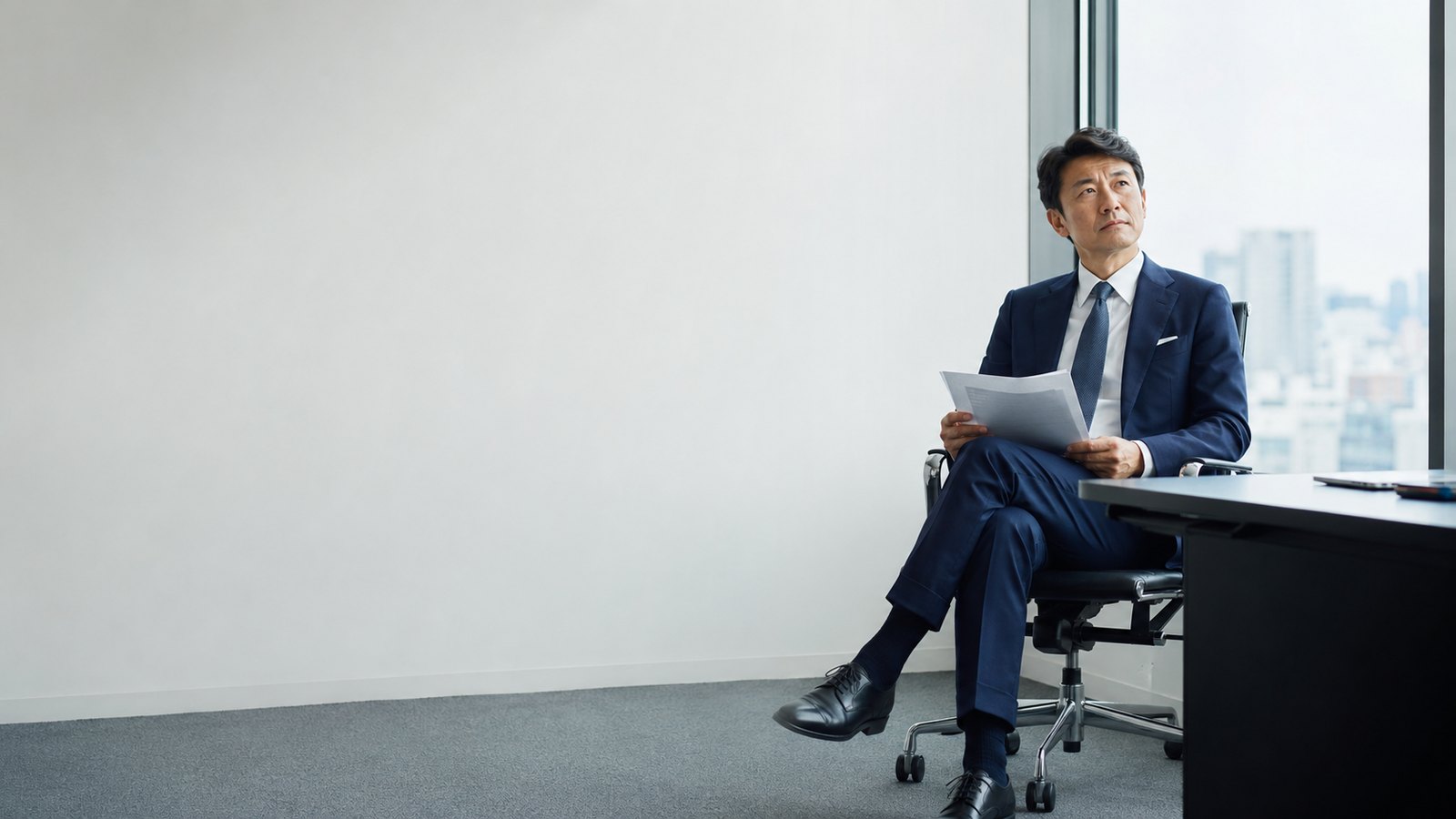 窓際の椅子に腰かけ、手元の書類から目を上げて考えている男性経営者